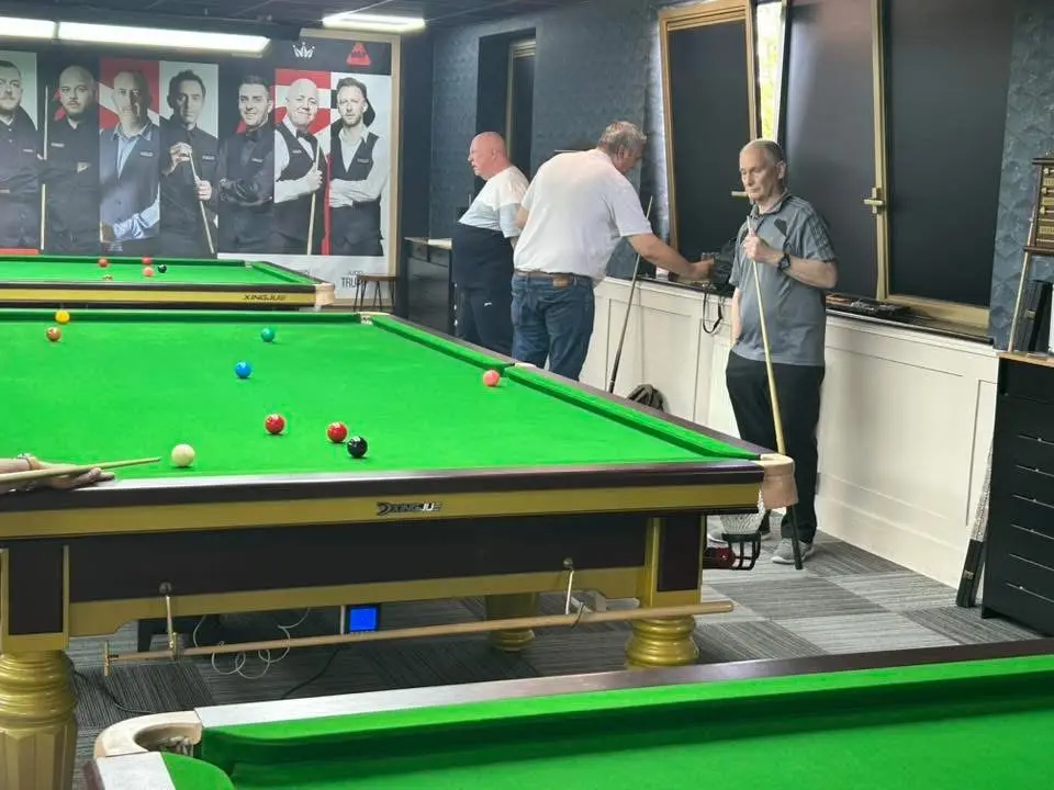 Players standing between tables in conversation, one waiting with his cue