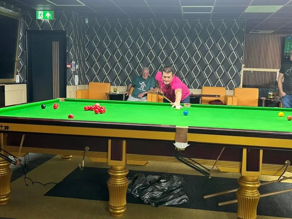 A player cueing along the cushion while an opponent watches from a chair
